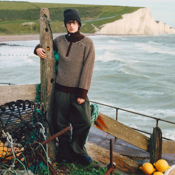 Person wearing round glasses by a windswept beach, leaning on a wooden post.