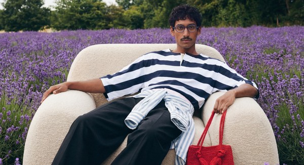 A person sits in a cream armchair in a lavender field, wearing a striped shirt and holding a red woven bag, looking ahea
