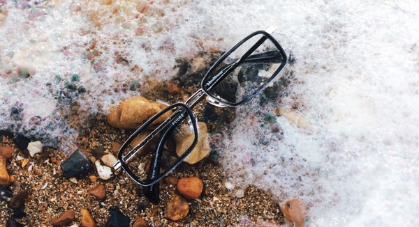 Rectangular eyeglass frames lying in sea foam on a pebbled beach.