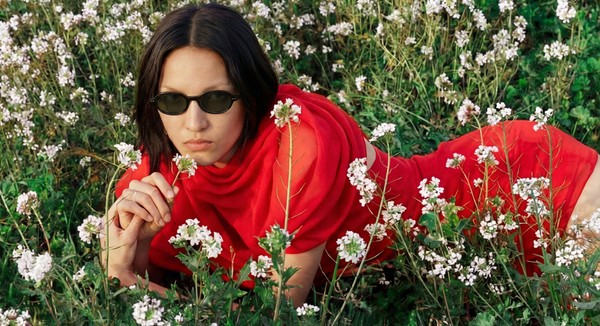 A person in a bright red outfit and narrow sunglasses lies among small white flowers, looking directly at the camera.