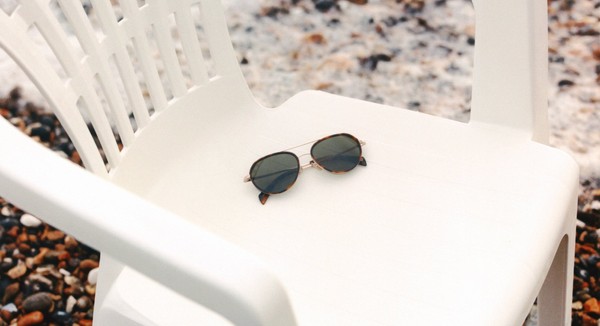 Round sunglasses placed on a white plastic chair on a pebbled beach.