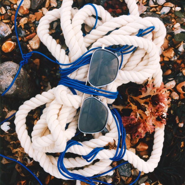 Sunglasses resting on thick white rope and blue cord on a pebbled beach.