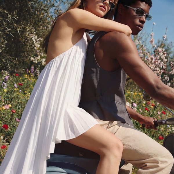 Two people wearing sunglasses ride a scooter through a flower-filled landscape; one in a white dress leans on the other.