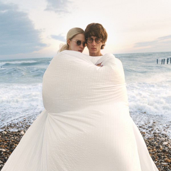 Two people wrapped in a white blanket on a pebble beach, both wearing glasses.