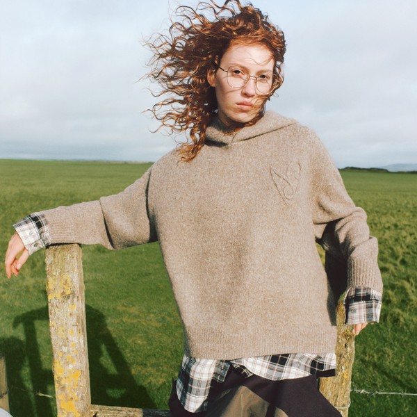 Model wearing eyeglasses outdoors, hair blowing across face in open countryside.