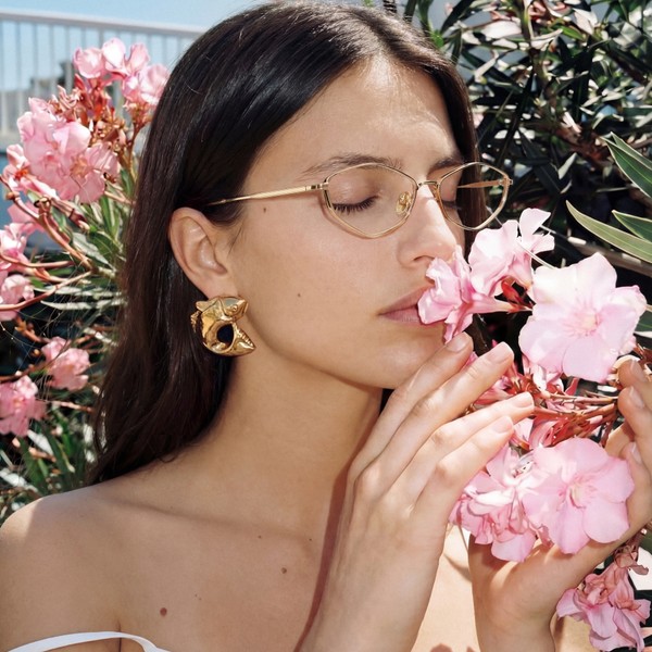 Close-up of a person wearing gold-framed glasses and sculptural gold earrings, gently smelling pink flowers with eyes cl