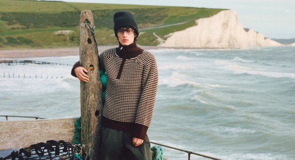 Model in glasses and knitwear posing at the seaside with waves behind.