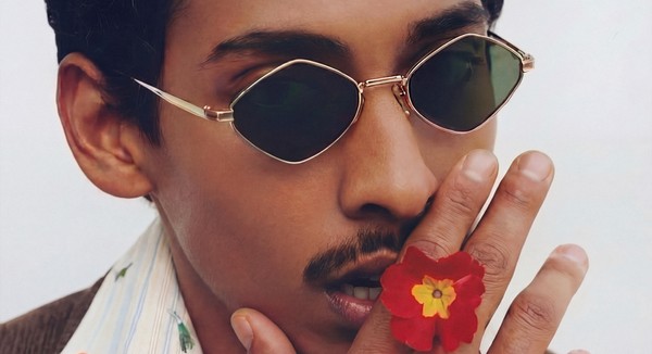 A person wearing sunglasses holds a red flower near their face, partially covering their mouth.