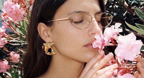 Close-up of a person wearing gold-framed glasses and sculptural gold earrings, gently smelling pink flowers with eyes cl