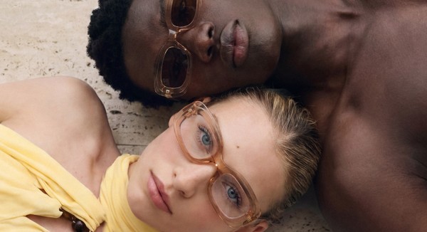 Two models wearing translucent blush square optical glasses while lying on a stone surface in the sun.