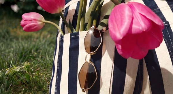 Gold-framed sunglasses hang from striped trousers beside pink tulips in a sunlit garden.