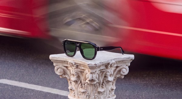 Black bloobloom sunglasses on a stone plinth as a red London bus passes behind.