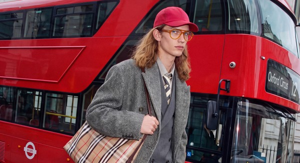Model in orange glasses and grey tailoring walking beside a red London bus.