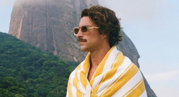 Male model wearing RIPPLE sunglasses wrapped in a yellow-striped towel against Rio's mountainous coastline.