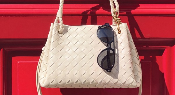 Black bloobloom sunglasses hanging from a cream woven handbag against a red background.