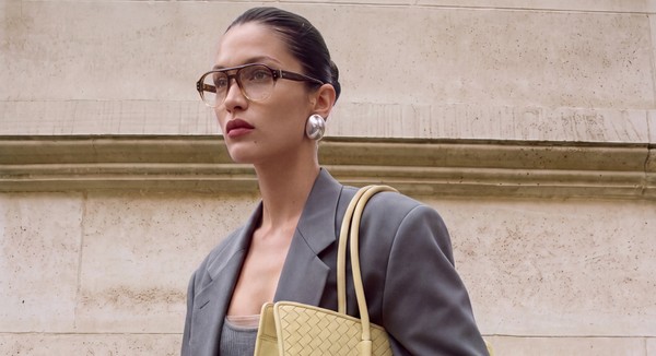 Model wearing brown glasses and grey tailoring with a woven yellow handbag.