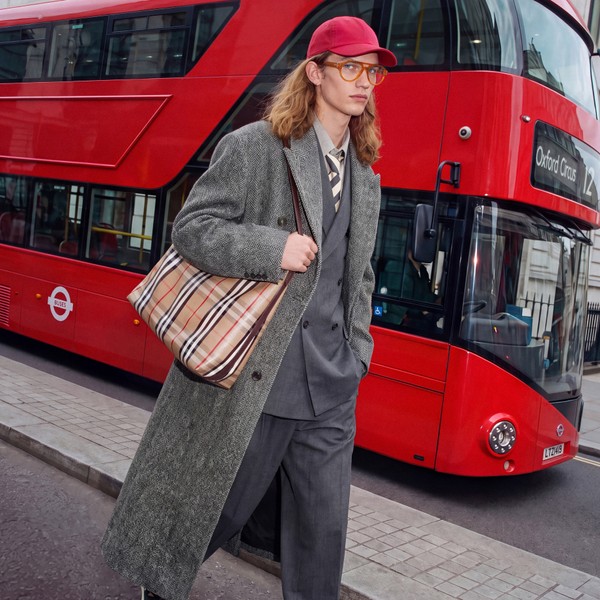 Model in orange glasses and grey tailoring walking beside a red London bus.